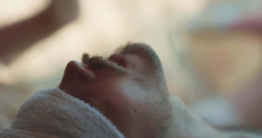 A man lying down removes a towel from around his neck and puts it over his eyes as a woman fans him