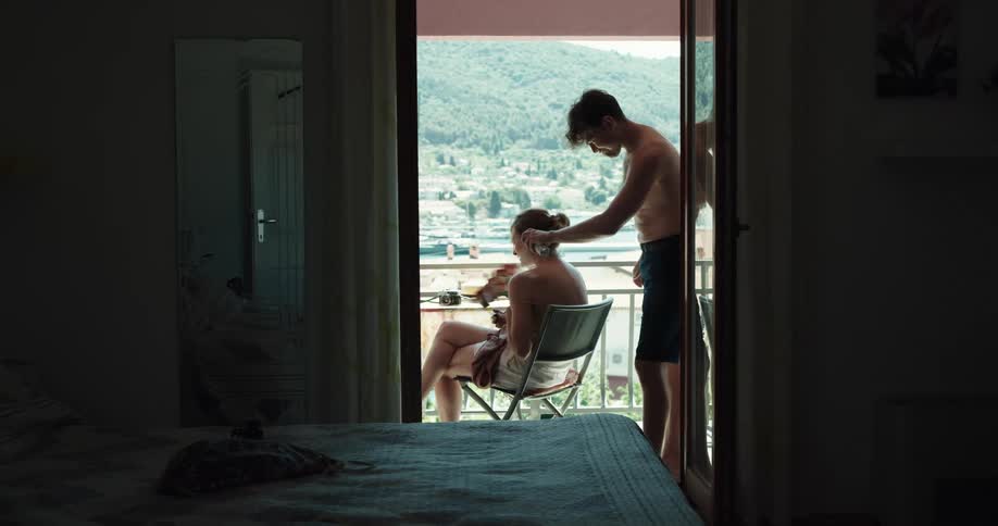 A woman sits on a balcony in a hotel as a man sprays sunscreen on her shoulders
