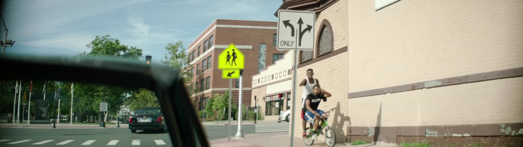 Two teens riding a bicycle down a sidewalk smile and point at a car going by