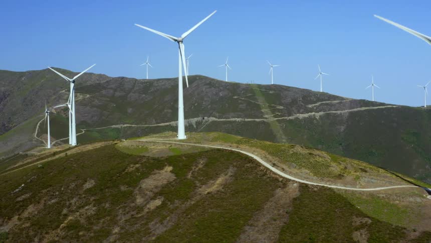 Road wines across top of hills and along mountain slope past wind turbines under clear blue sky