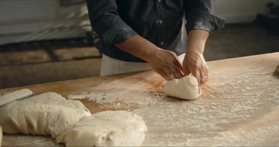 A man in a room kneading dough on a table with flour with a wall in the background