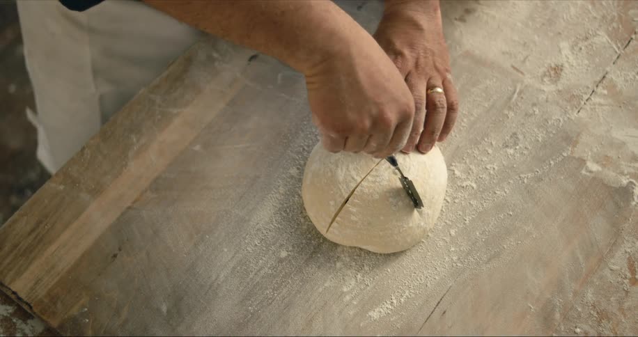 Man with black shirt and white apron scores dough with bread lame on wood