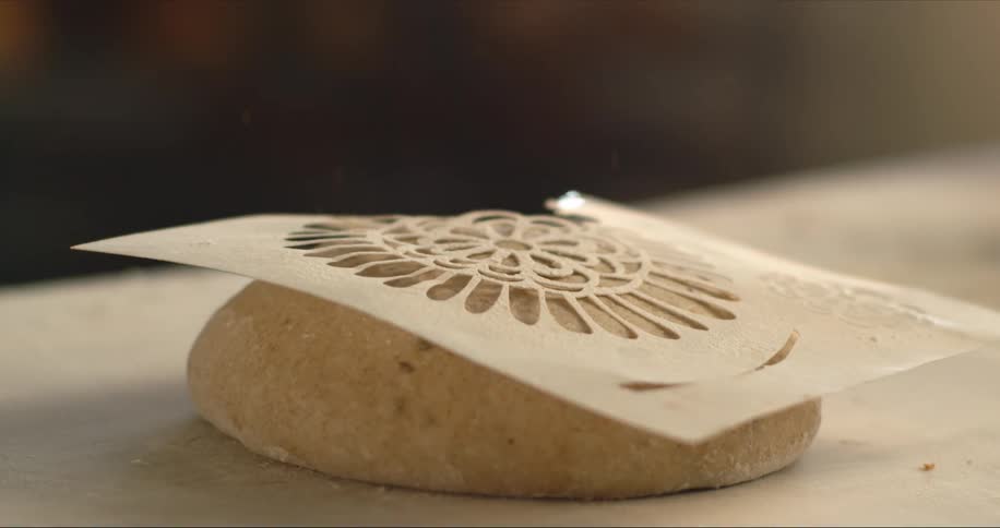 A person lays a stencil on some dough then sprinkles flour on top
