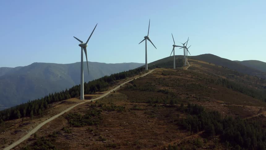 Mist obscures mountain in background as row of wind turbines rotate along side of road passing over mountain near forest