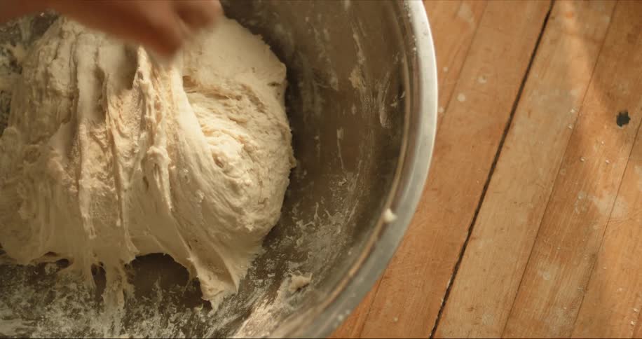 Man kneading bread dough inside a metallic bowl over a table
