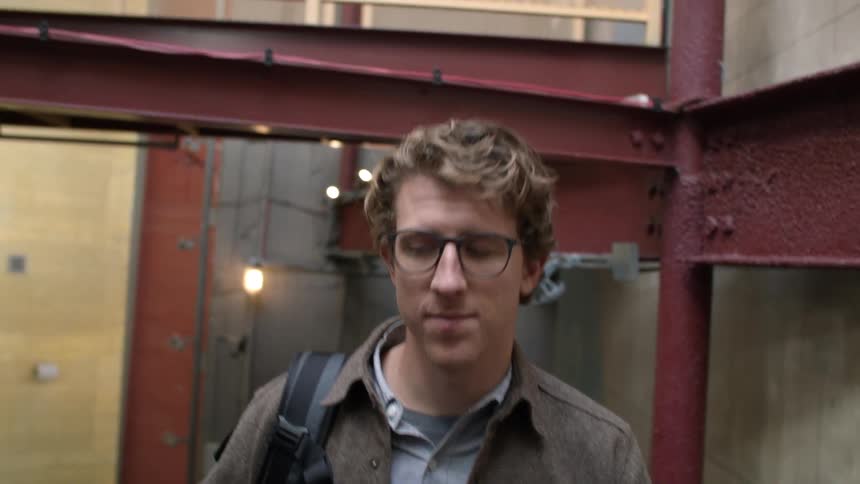 A man wearing a brown jacket and glasses is looking around as he is walking up stairs inside a building