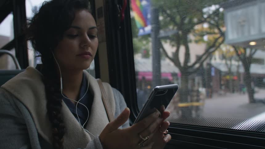 Woman with hair in plaits and earphones uses smartphone on moving bus