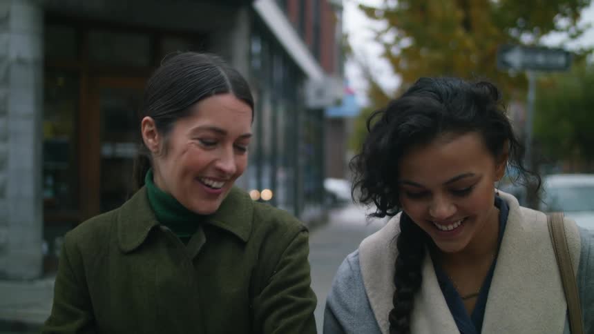 Two women are walking down a sidewalk talking and smiling as they pass buildings