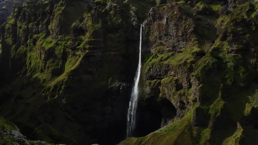 Aerial view of woman with black sweater and jeans standing on the edge of high cliff looking at high mountains with waterfall cascading down the mountainside