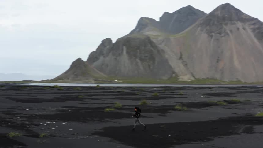 A woman is running across land with few plants and mountains in the nearby distance