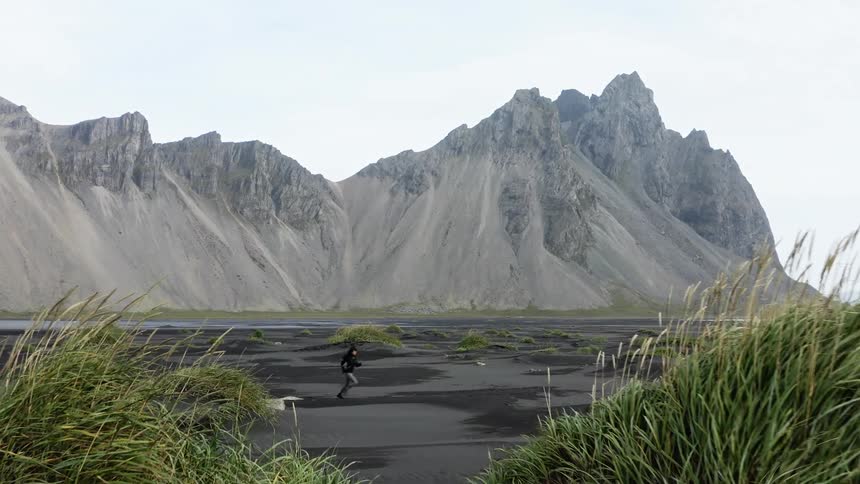 Woman jogs across black sand dotted with sea grass across water from mountain