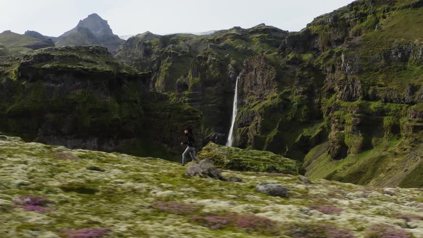 A woman running in a landscape with cliffs a waterfall the sky and clouds in the background