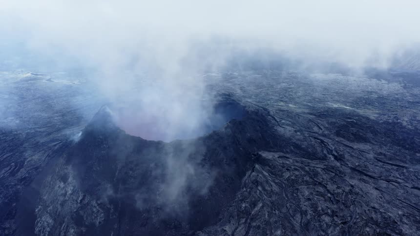 Clouds flow obscuring volcano surrounded by dark ash terrain