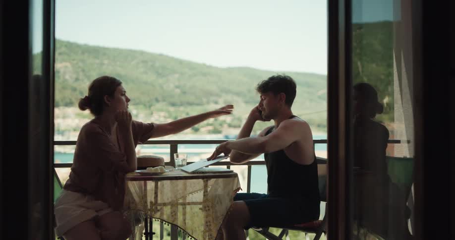Couple fight as woman tries to keep man from working while they sit on balcony overlooking water
