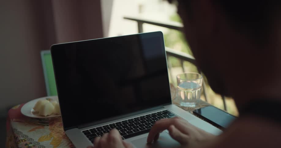 Man sitting near a balcony working on a laptop answers a phone call