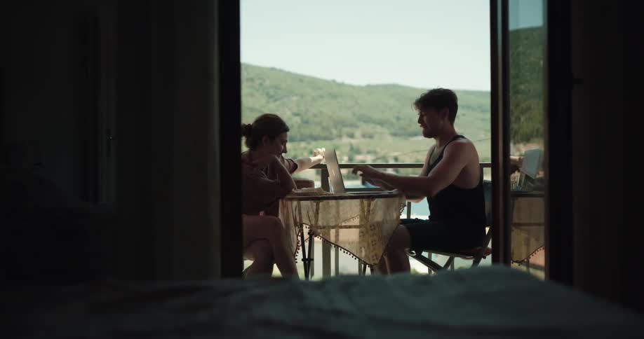 Man with black undershirt uses laptop and woman comes to take him joking with one another on the balcony