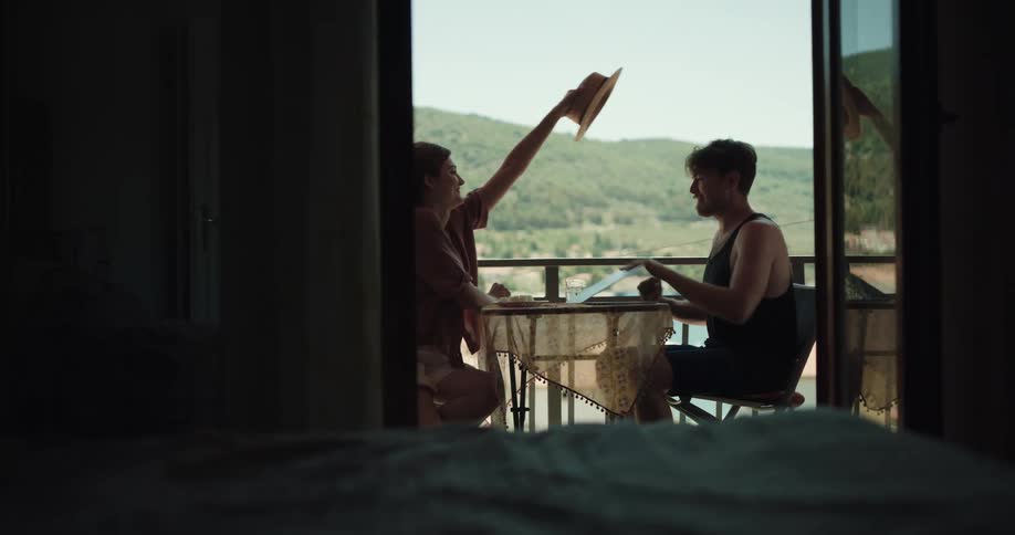 A man sits on a hotel balcony typing on a laptop when a woman comes out and puts a hat on his head and he closes his laptop takes a drink and grabs her by the hand and they leave
