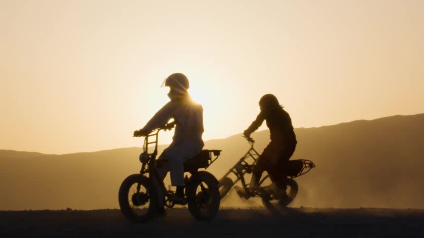 Two people are riding motor bikes in the dirt in the wilderness at sunset