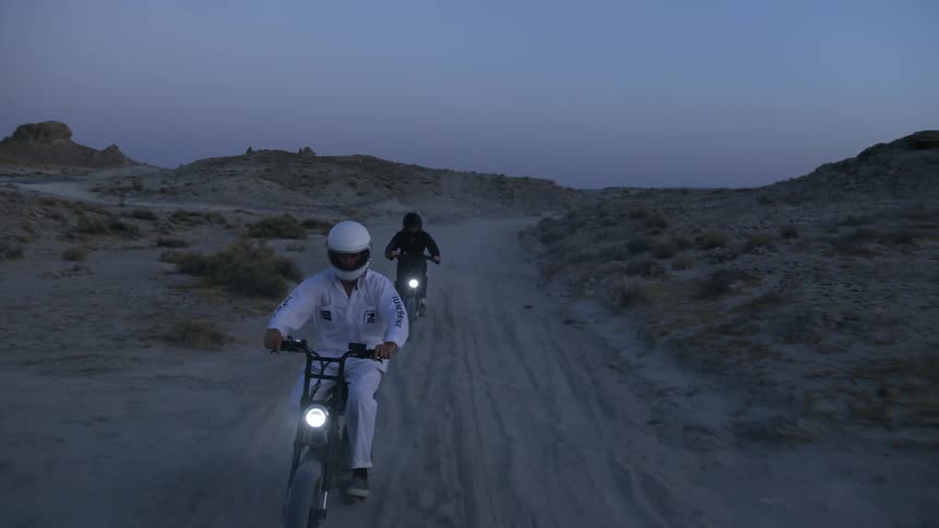 Two people riding motorcycles on a dirt road in a desert landscape with the sky and clouds in the background