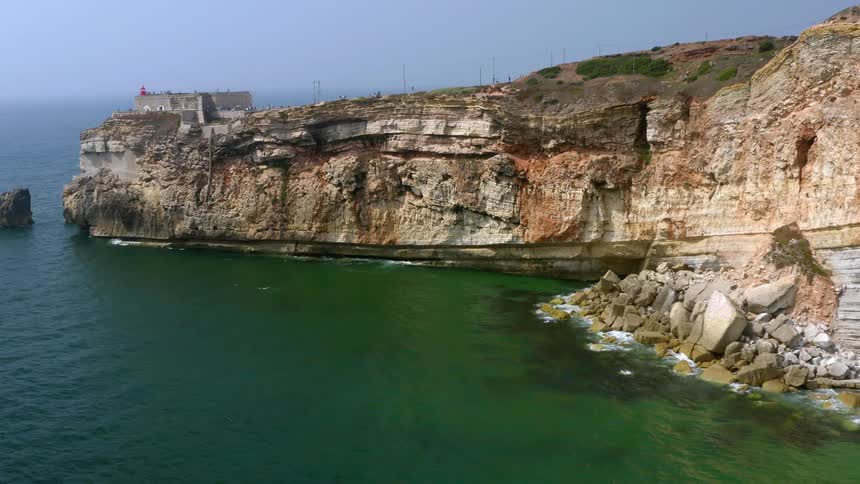 A rocky cliff stretches out into the sea with a building on the edge