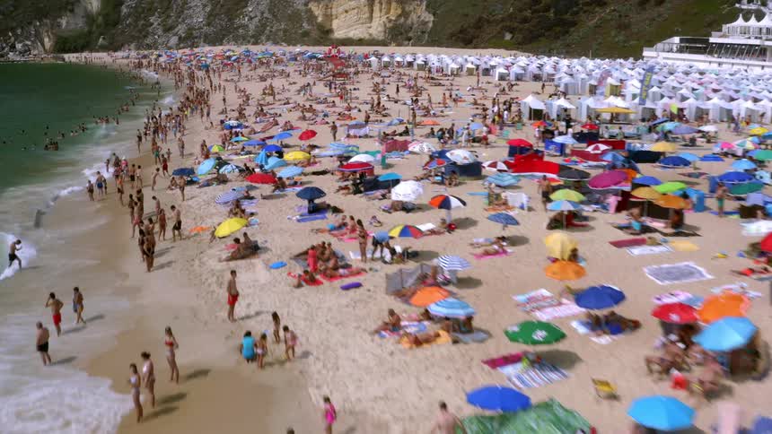 A sandy ocean beach is covered with crowds of people and umbrellas on a sunny day