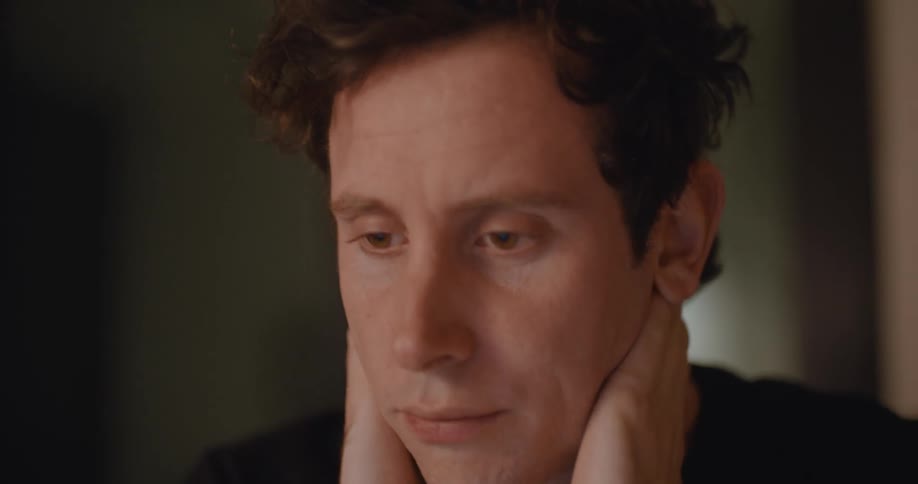 A man sitting at a desk rubs his face and neck tiredly as he looks down at his notes and laptop