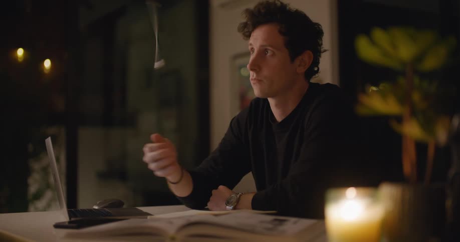 A man in a room sitting at a table next to a laptop playing with a pencil with a wall in the background
