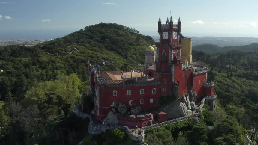 An aerial view of a castle surrounded by a forest atop a mountain ridge with a town in the valley below