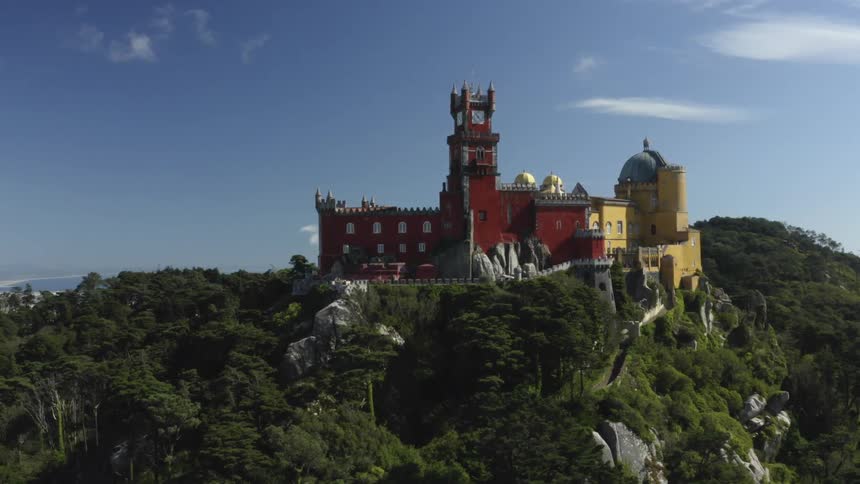 A multicolored castle sits on top of a tree covered cliff