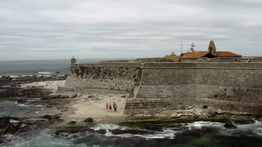 A stone wall surrounds a building along the coastline with mountains in the distance