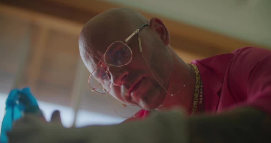 A man in a room with glasses and gloves giving a tattoo to someone with a window in the background