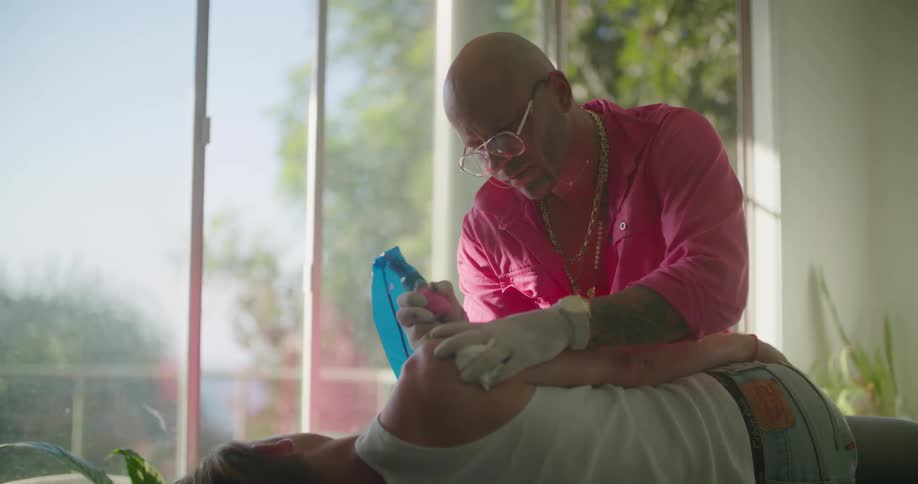 A man in a room with gloves giving a tattoo to another man with a wall and window in the background
