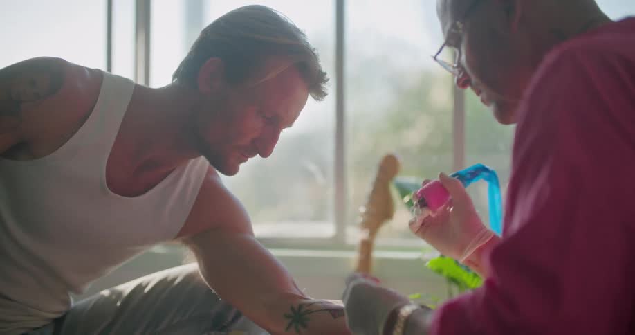 A man with glasses and gloves in a room giving a tattoo to another man with a window in the background