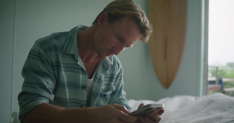 A man uses his smartphone while sitting on a bed in a bedroom during the day
