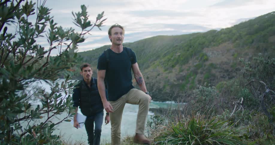 Two men with a backpack and notepad are walking up a hill overlooking the ocean