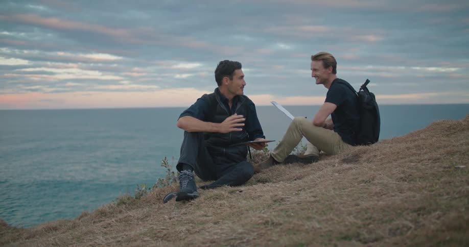 A pair of men sitting on a hill overlooking the ocean talk as one uses a computer tablet and the other writes on a clipboard