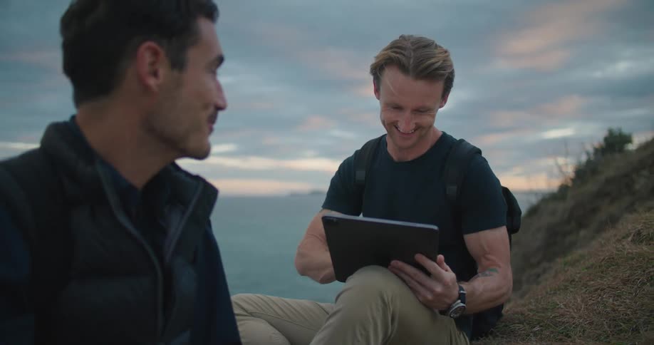 Two men are sitting and talking on a grass covered hill along the shore and one of the men is holding a computer tablet and the other is writing on paper