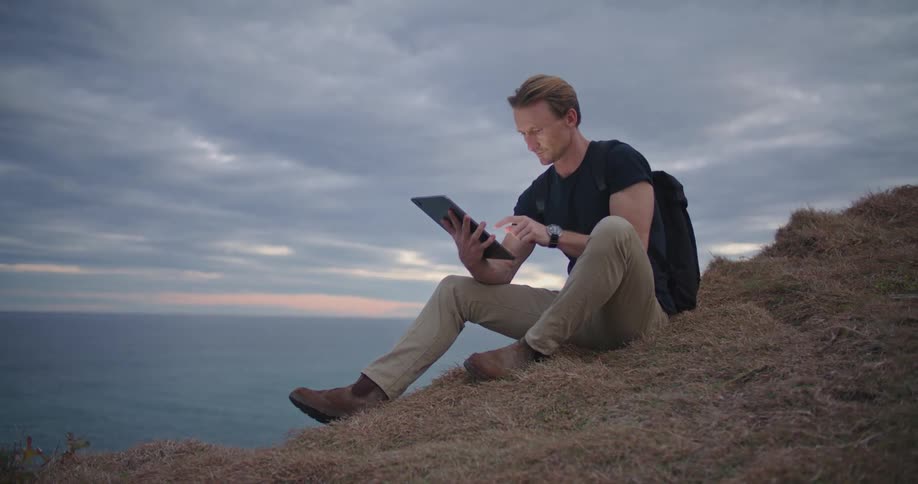 A man with a tablet and a backpack sitting on a cliff with the ocean the sky and clouds in the background
