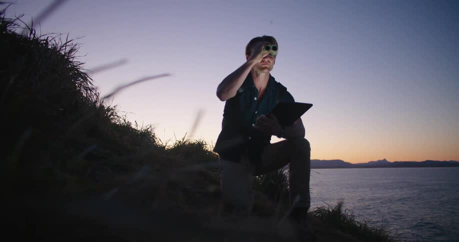 A man is sitting in the grass on a hill along the shore holding a computer tablet in one hand and binoculars in the other hand