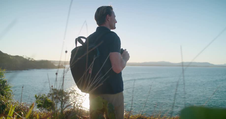 A man with a backpack standing in front of a lake looking through binoculars with the sky and clouds in the background