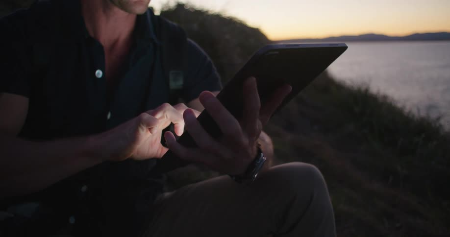 A man sitting on the shore of a lake with a tablet raises a pair of binoculars to his face to look out over the lake