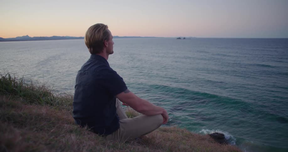 A man is sitting in the grass on a hill watching ocean waves rolling in