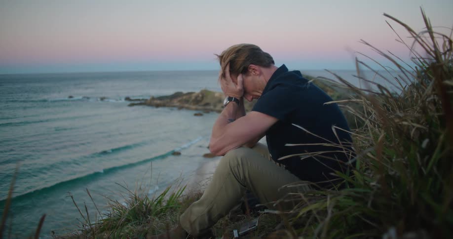 A man rubs his face while sitting on a hill overlooking the ocean