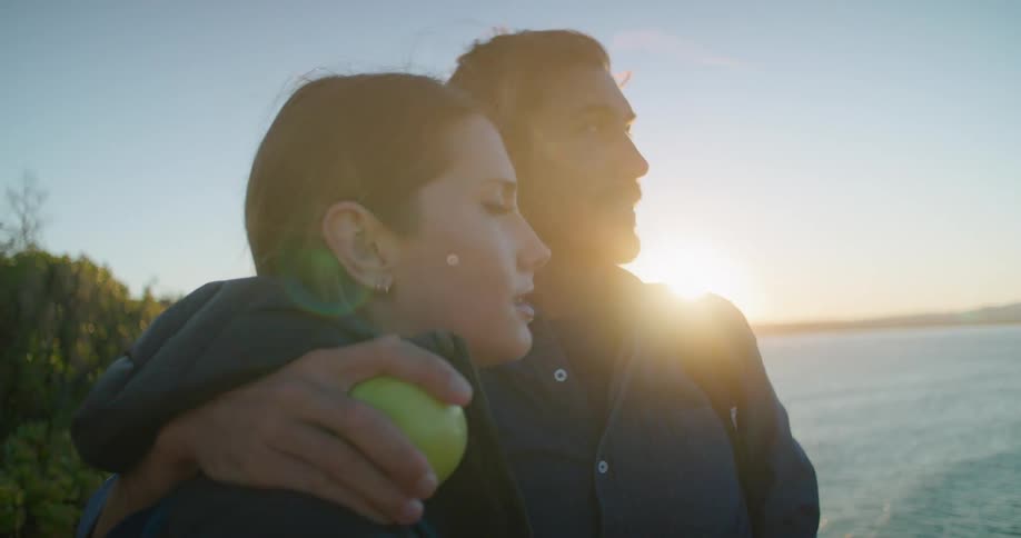 A man stands with his arm around a woman on the shore of a lake as they look out and talk about their surroundings