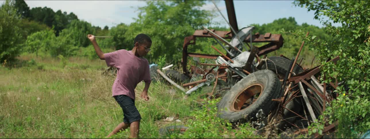 Boy swings stick at plant before climbing pile on metal scraps