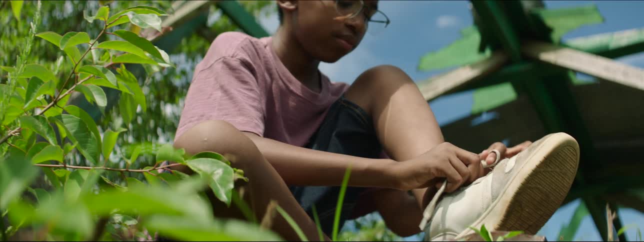A boy is sitting down near plants and holding a stick then he lets go of the stick to tie his shoe