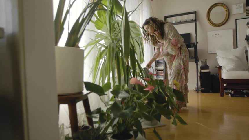 A pregnant woman in a room watering plants with a water can with a wall and window in the background