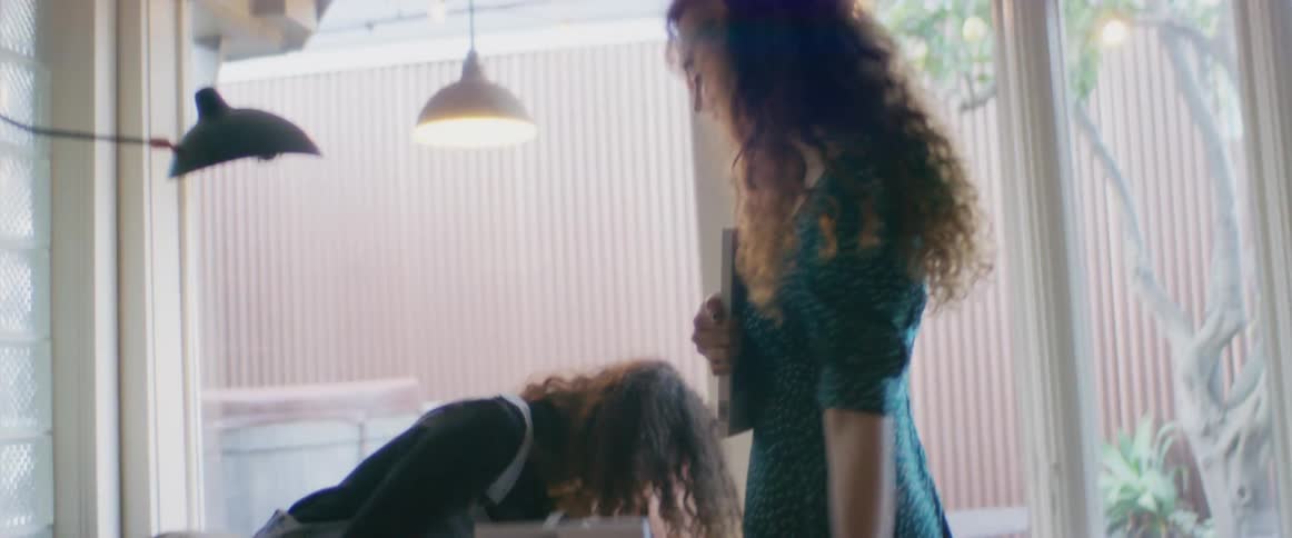 Two women in a room with one holding a smart phone sitting on a bed next to a bookshelf with a window in the background