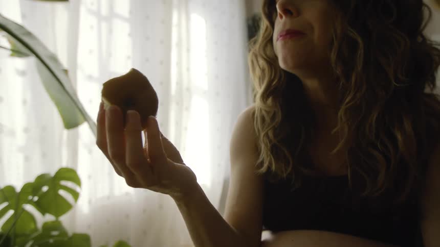 A pregnant woman is sitting indoors eating a pear near a window during the daytime