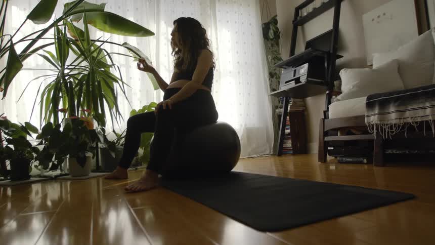 A pregnant woman eats while sitting atop an exercise ball in the living room of a house during the day